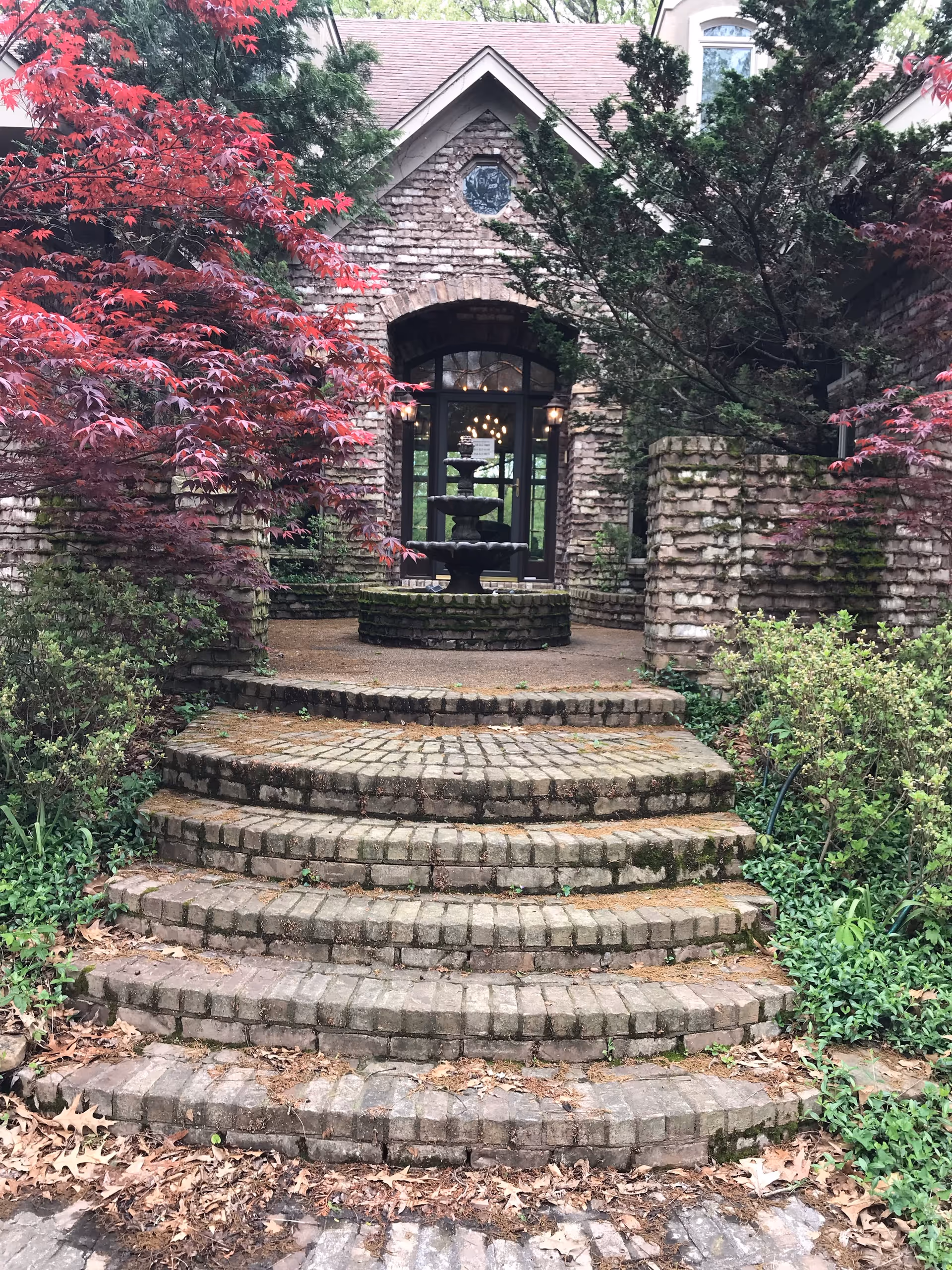 Stone steps leading up to a stone building entrance with a multi-tiered black fountain in front. The area is surrounded by lush greenery and trees with red and green leaves.