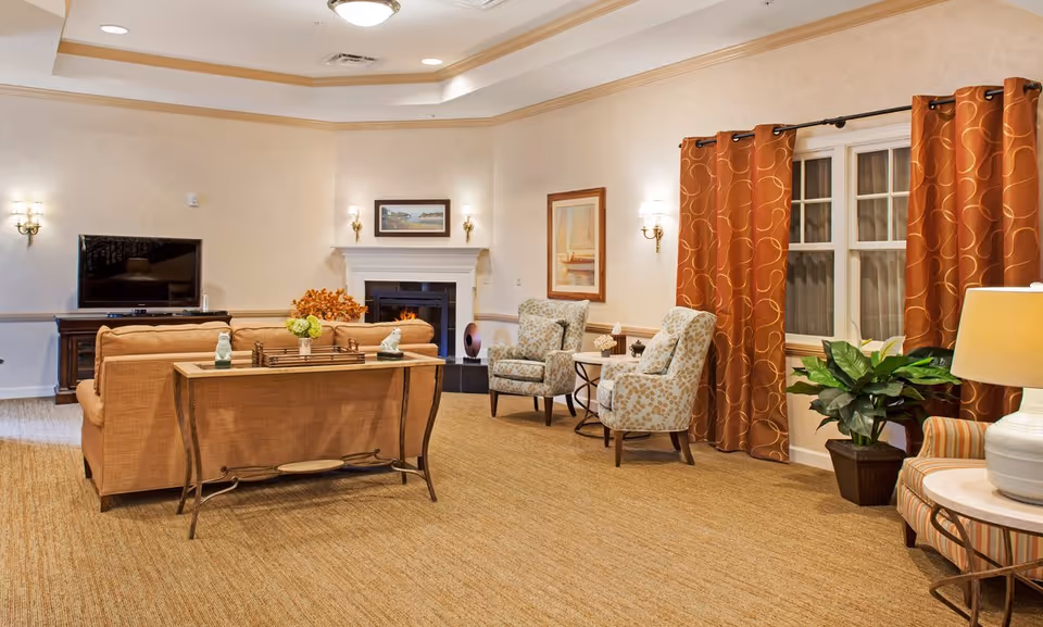 A cozy living room area with a beige couch, two patterned armchairs, a wooden console table, and a fireplace with a framed picture above it. There is a flat-screen TV on a wooden stand to the left, orange curtains with a swirl pattern on the windows to the right, and a potted plant near a round side table with a lamp.