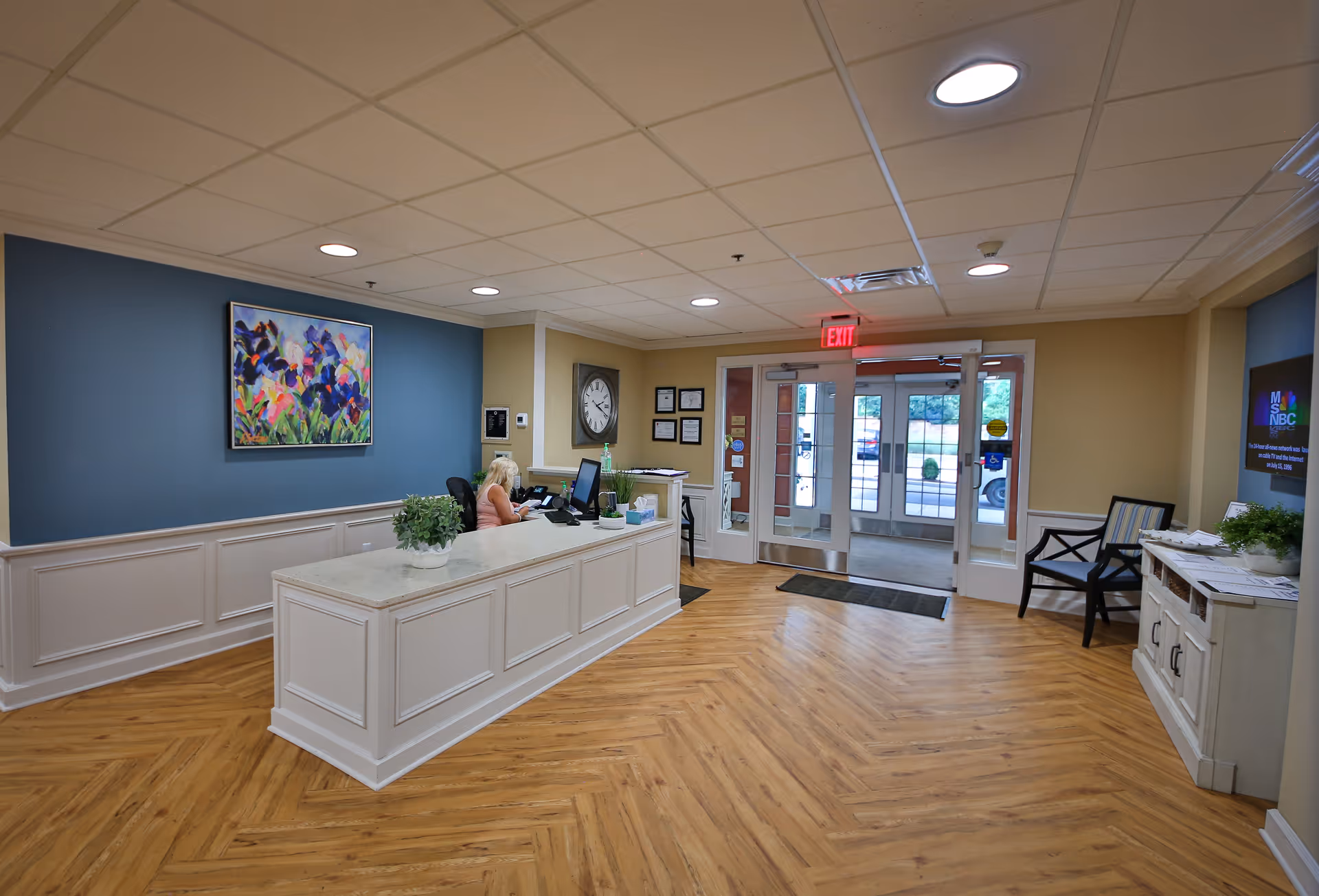 Reception area with a front desk, a receptionist, seating and glass entrance doors in an assisted living facility.