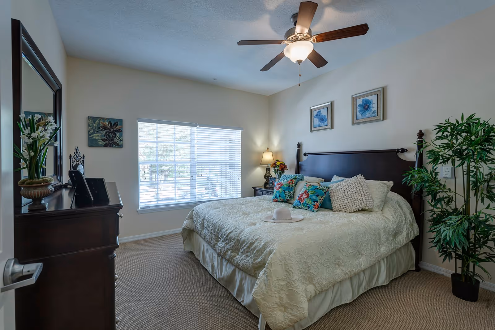 A cozy bedroom featuring a large bed with decorative pillows, a ceiling fan, and a window with blinds. A dresser and a potted plant are visible, creating a warm and inviting atmosphere.