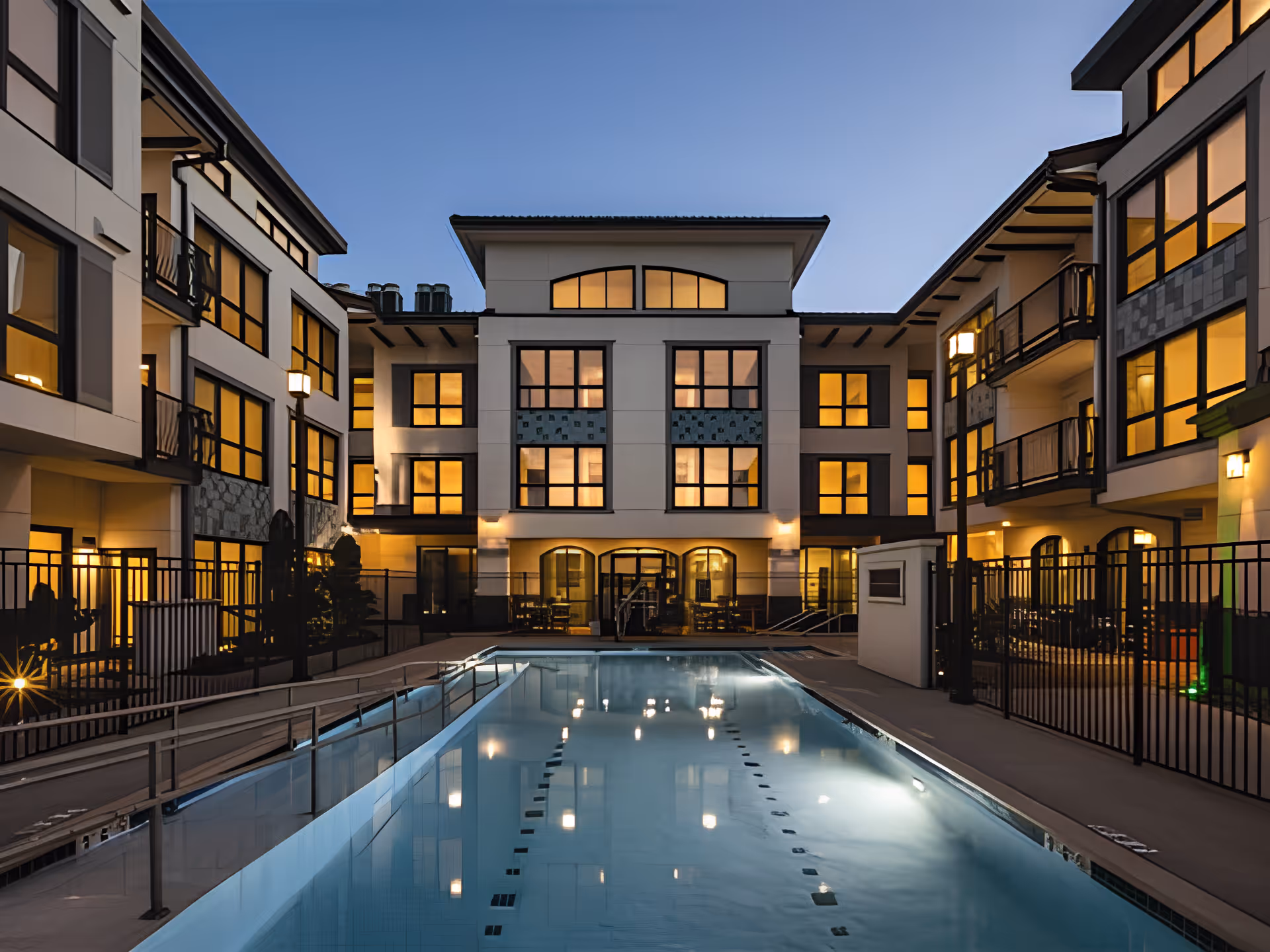 A lit courtyard swimming pool at dusk surrounded by a three-story senior living building with balconies and illuminated windows.