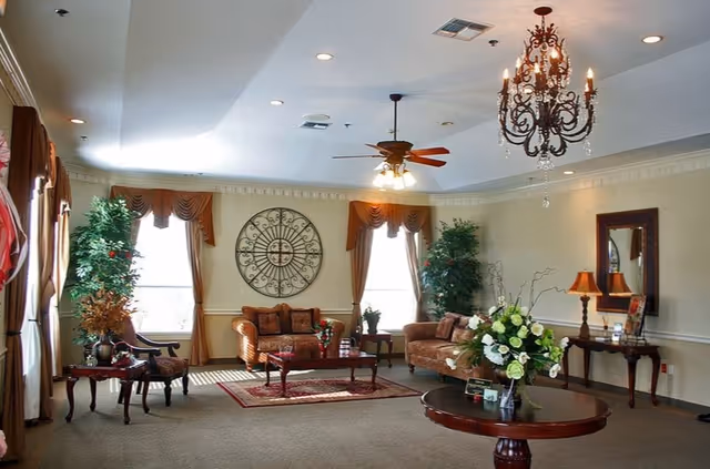 A well-lit living room with two large windows draped with brown curtains. The room features two brown upholstered sofas, a wooden coffee table on a red patterned rug, a round wooden table with a floral arrangement, and two potted plants near the windows. There is a decorative wall piece between the windows, a ceiling fan with lights, and a chandelier hanging from the ceiling. A wooden console table with a lamp and a mirror is against the right wall.