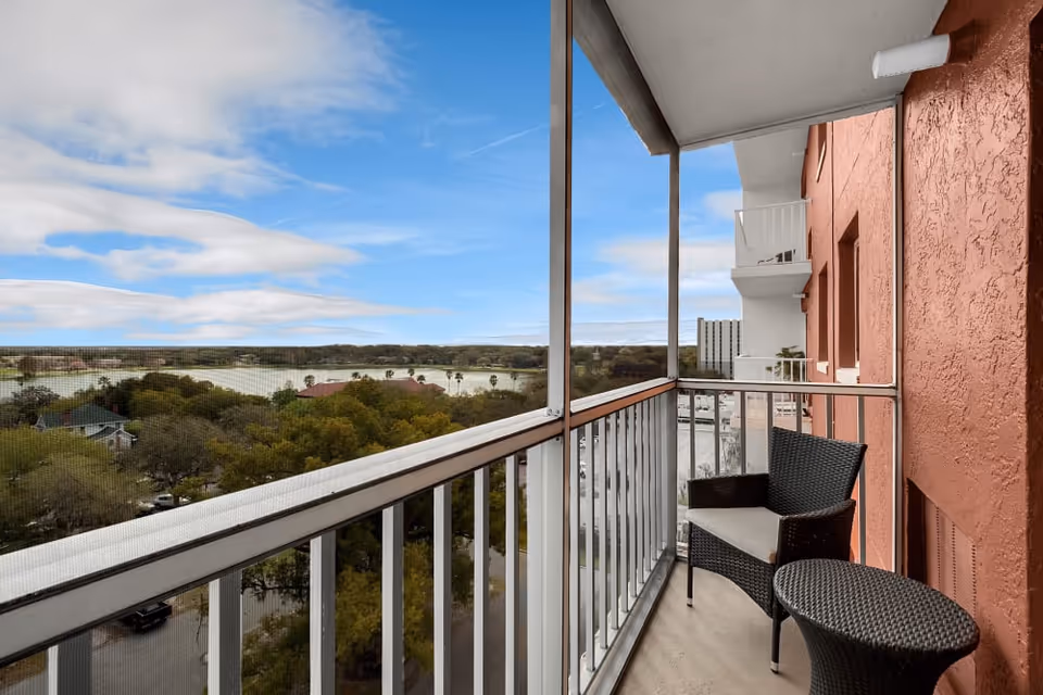 View from a screened balcony with a black wicker chair and matching small table, overlooking trees, a lake, and a partly cloudy sky.