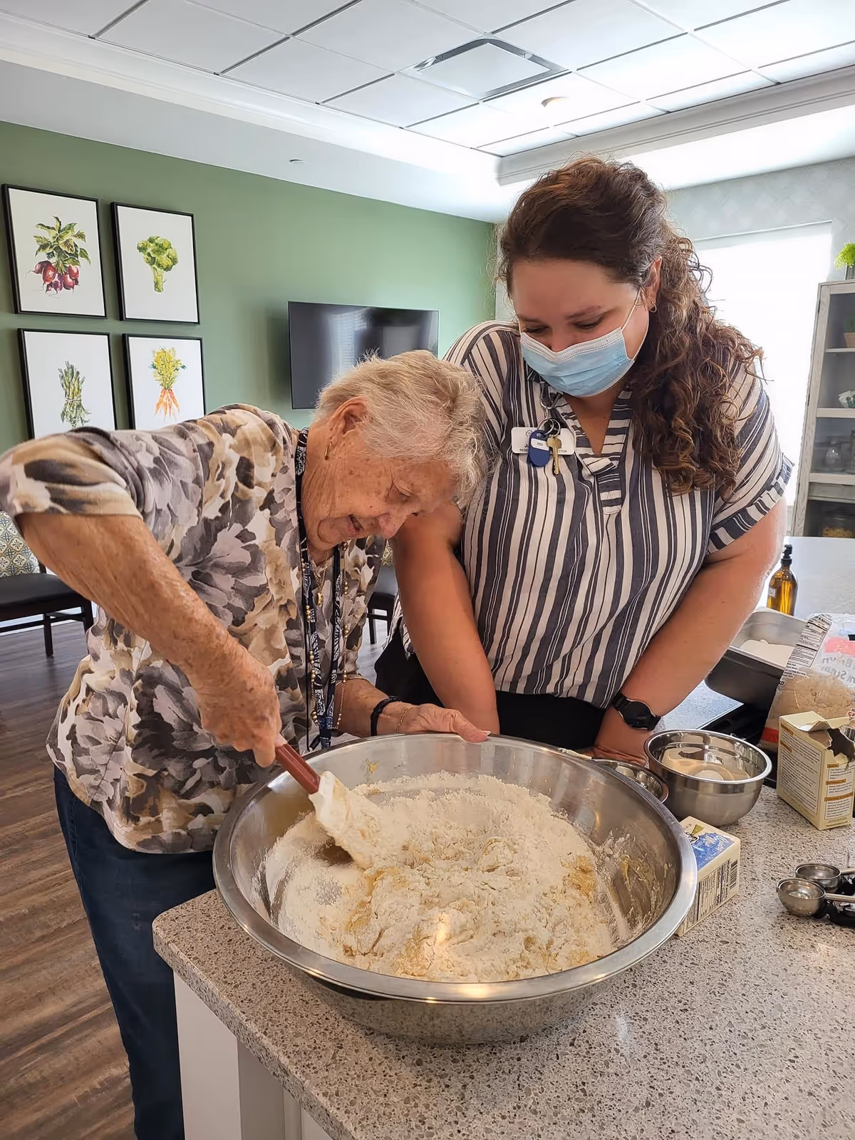 An elderly woman stirring a large bowl of flour mixture on a kitchen counter while a younger woman wearing a face mask watches and smiles beside her. The setting appears to be a bright, modern kitchen area with green walls and framed botanical prints in the background.