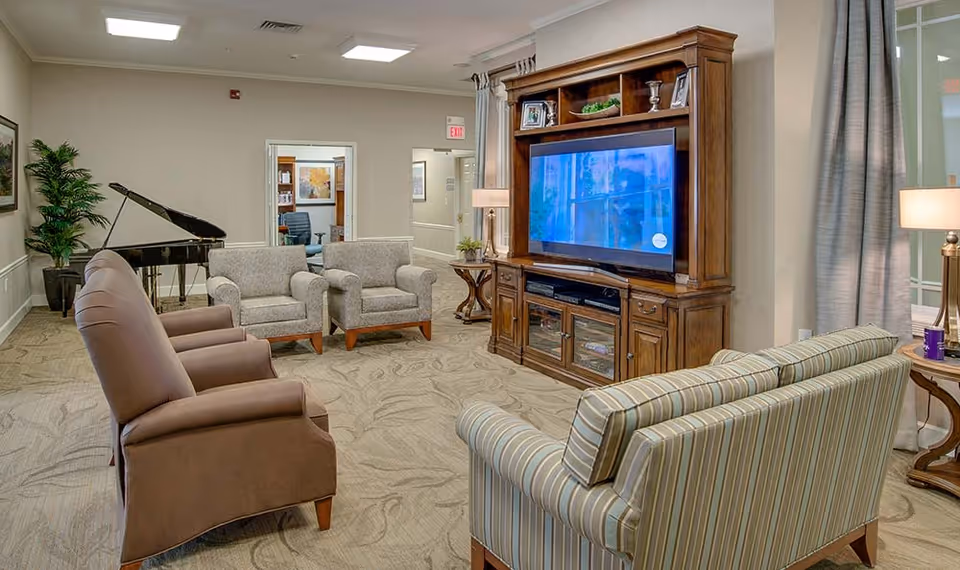 A senior living facility common area with a large wooden entertainment center holding a flat-screen TV, surrounded by various seating options including a striped sofa, two beige armchairs, and two brown armchairs. There is a grand piano in the corner and a potted plant next to it. The room has beige walls, carpeted floor, and ceiling lights.