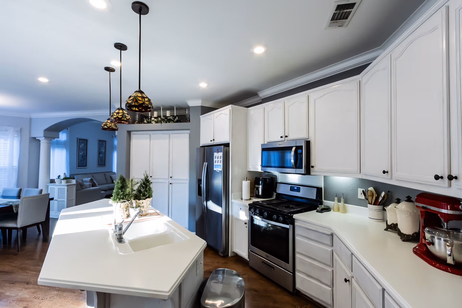 Bright modern kitchen with white cabinets, a large white island with a sink, stainless steel appliances including a refrigerator, stove, and microwave. Three pendant lights hang above the island. The kitchen opens into a dining and living area with a table and chairs visible.