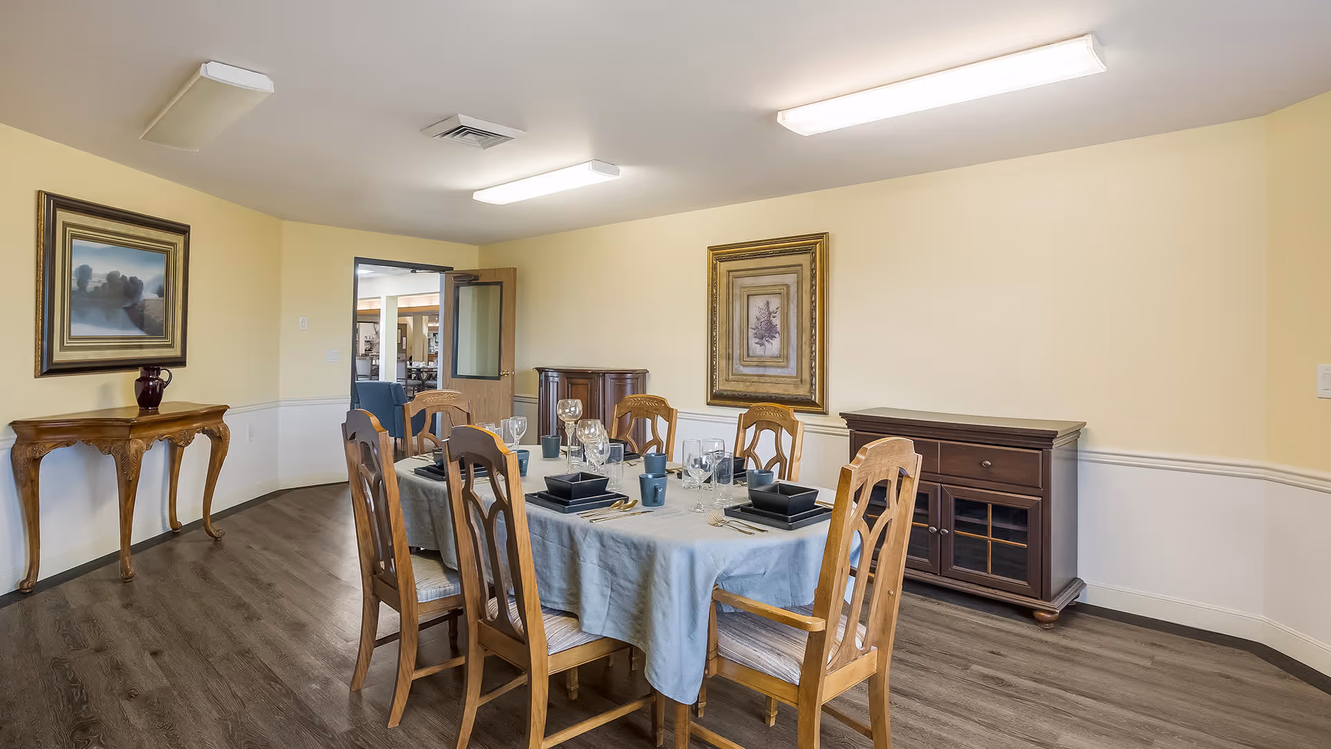 A dining room with a rectangular table covered with a light blue tablecloth, set with black square plates, glasses, and silverware. Six wooden chairs surround the table. The room has light yellow walls with white wainscoting, two framed paintings, a wooden sideboard, and a small wooden console table with a decorative vase. The floor is a dark wood laminate, and there is a door leading to another room in the background.
