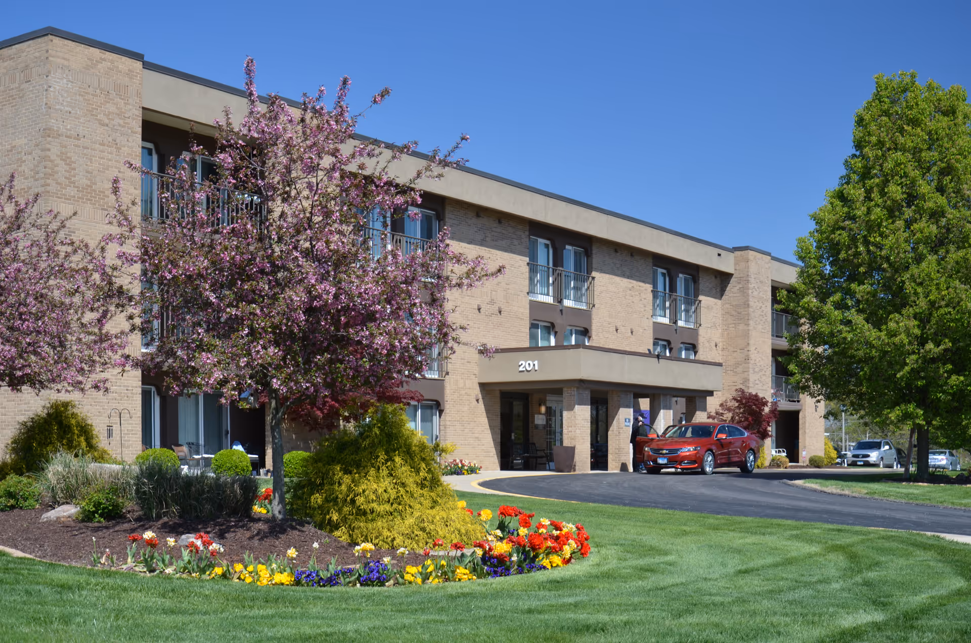 Front exterior of a three-story brick senior living building with a manicured lawn, flower beds, blossoming trees, and cars parked at the entrance marked 201.