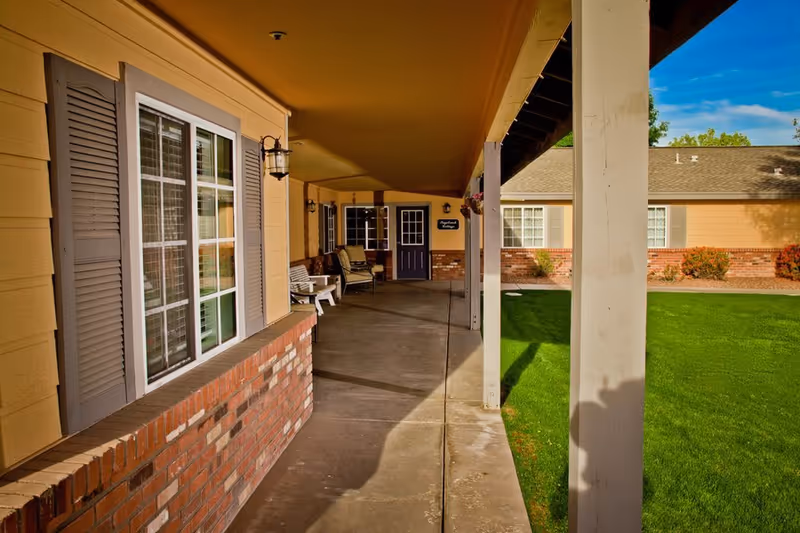 Covered walkway outside a senior living facility with brick and yellow siding walls, windows with shutters, outdoor seating, and a well-maintained green lawn under a blue sky.