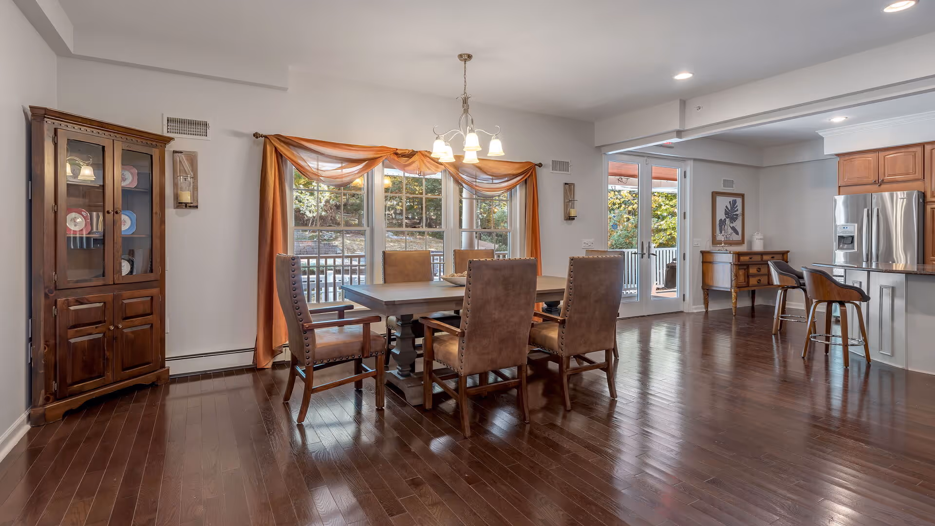 A spacious dining area with a wooden dining table surrounded by six upholstered chairs. To the left, there is a wooden cabinet with glass doors displaying plates. Large windows with orange curtains let in natural light. In the background, there is a kitchen area with wooden cabinets, a stainless steel refrigerator, and a breakfast bar with two stools. The floor is dark hardwood, and the walls are painted light gray.