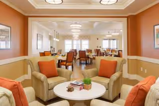 A cozy common area with beige armchairs and orange cushions arranged around a round white table with books and a small plant. In the background, there is a spacious dining area with multiple tables and chairs, large windows letting in natural light, and warm-toned walls.
