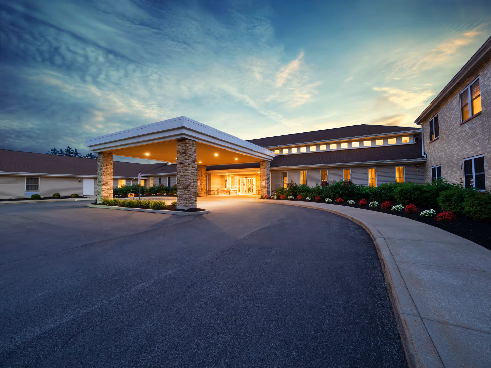 Exterior view of Brompton Heights of Williamsville senior living facility at dusk, showing the entrance with a covered drop-off area supported by stone pillars, well-lit windows, and landscaped flower beds along the sidewalk.