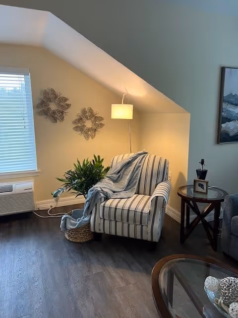 A cozy corner of a living room with a striped armchair draped with a light blue blanket, a potted plant in a woven basket on the floor, a floor lamp providing warm light, decorative wall art with three metal flower designs, a side table with a framed photo and a small decorative item, and a window with blinds partially open.