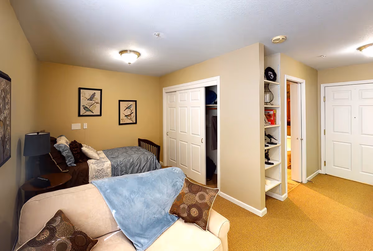 A cozy bedroom with a single bed against a beige wall, decorated with two framed bird pictures. Next to the bed is a small round table with a lamp. In the foreground, there is a beige couch with patterned pillows and a blue throw blanket. The room has a closet with sliding white doors and an open shelving unit with various items. The floor is carpeted in a warm brown color, and there are two white doors visible, one leading to another room and one likely an entrance door.