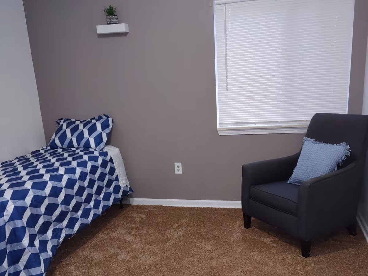 A simple bedroom with a single bed covered in blue and white geometric patterned bedding, a gray armchair with a light blue pillow, a small white floating shelf with a small plant on the wall, and a window with closed white blinds.