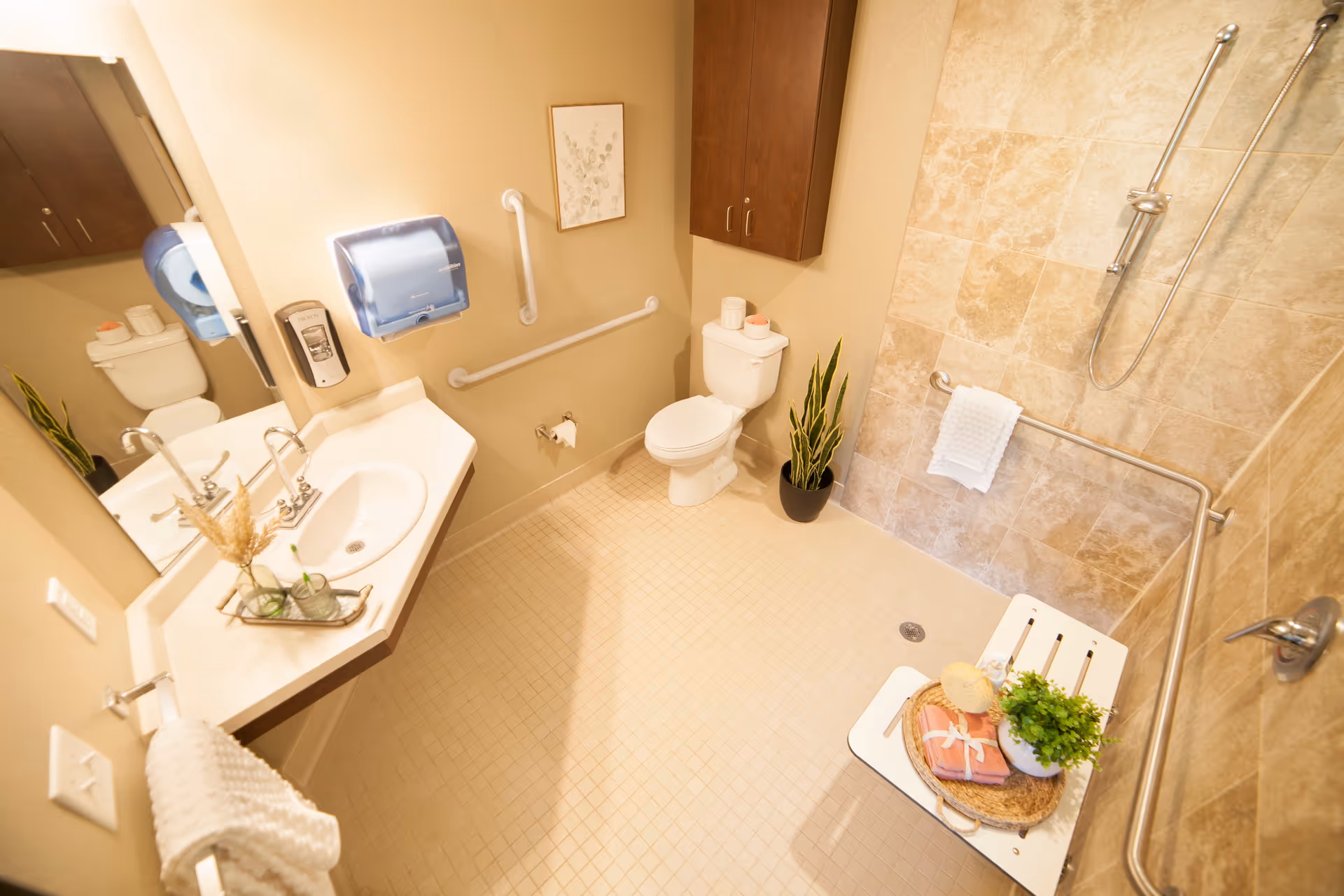 A spacious, accessible bathroom with beige tiled floors and walls. It features a white toilet, a wall-mounted sink with a mirror above, a blue paper towel dispenser, and a soap dispenser. There are grab bars near the toilet and in the shower area. The shower has a handheld showerhead, a white fold-down seat, and a towel hanging on a grab bar. The bathroom is decorated with a small plant near the toilet, a framed picture on the wall, and a tray with towels and a plant on the shower seat.