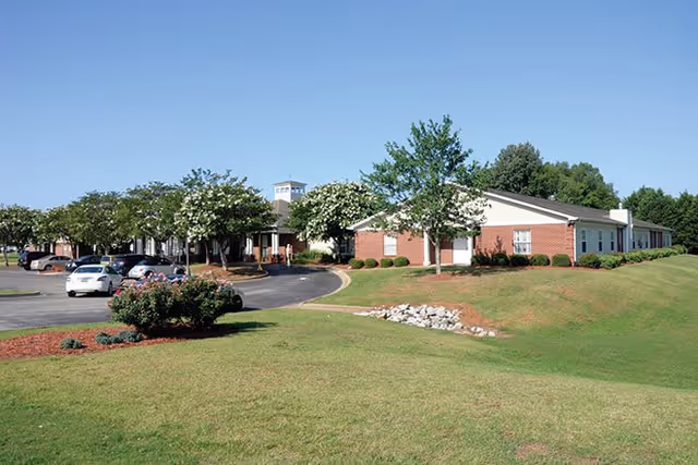Exterior view of a single-story brick building with a white roof, surrounded by green grass, trees, and a parking lot with several cars under a clear blue sky.