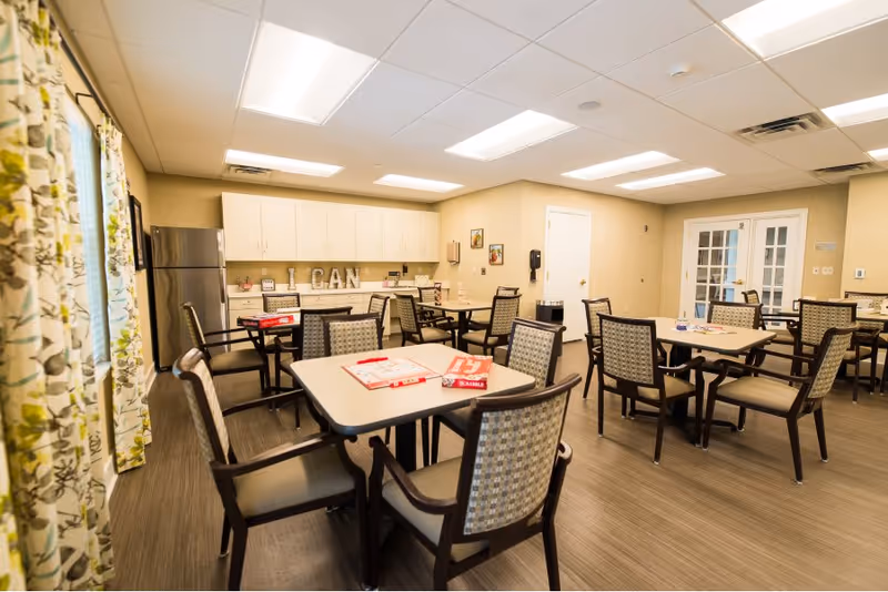 A bright and spacious room with multiple tables and chairs arranged for group activities or dining. The room features a kitchenette area with white cabinets and a stainless steel refrigerator. Board games are placed on the tables, and the walls are decorated with framed pictures and a sign that reads 'I CAN'. Large windows with floral curtains allow natural light to enter the room.