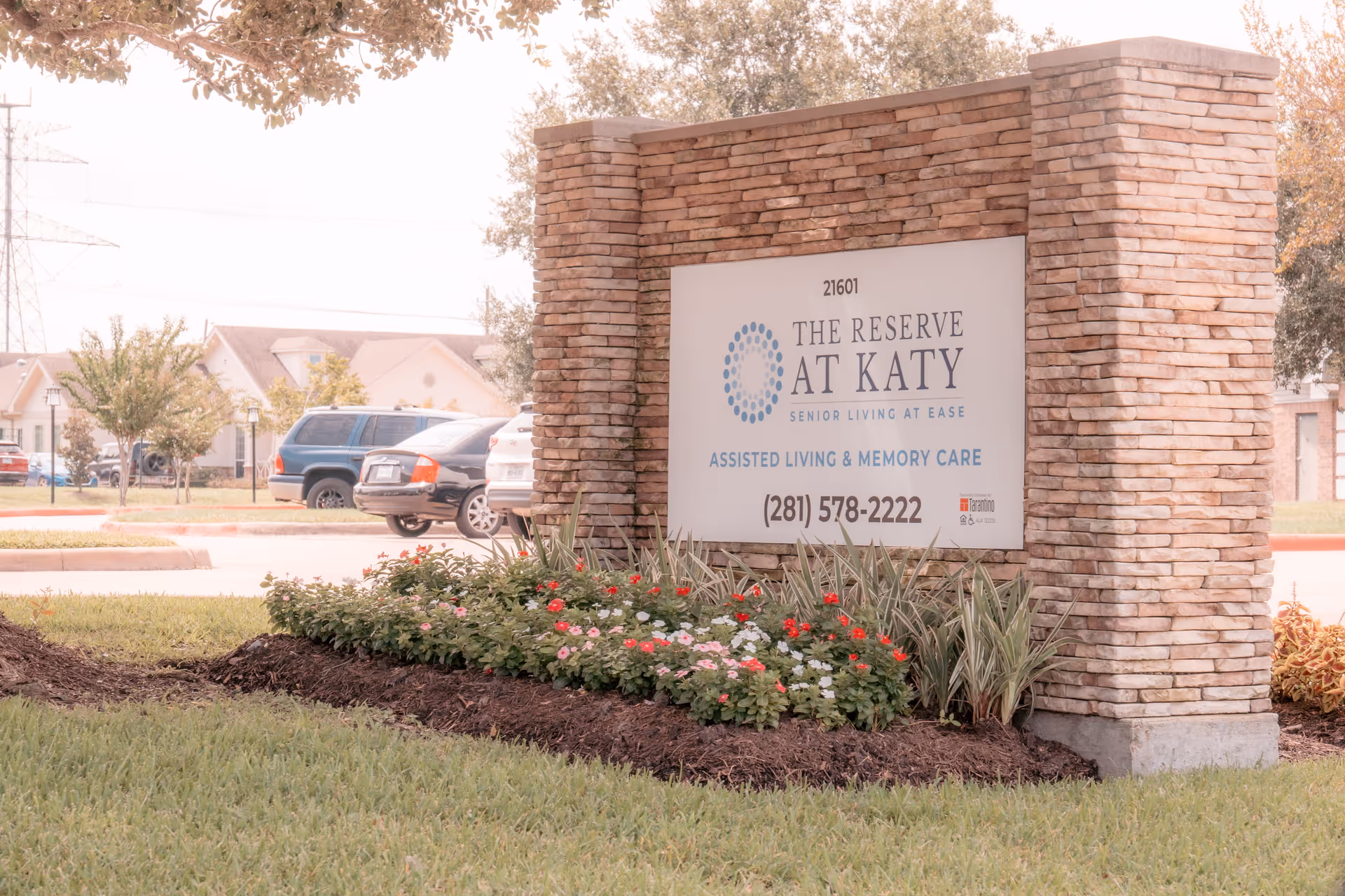 Stone monument sign reading 'The Reserve at Katy Assisted Living & Memory Care' at the facility entrance with landscaping and parked cars behind it.