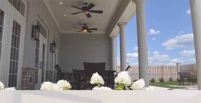 Covered outdoor patio area with ceiling fans, black chairs and tables, white columns, and white flowers in the foreground. A wooden fence and a bench are visible in the background under a blue sky with scattered clouds.