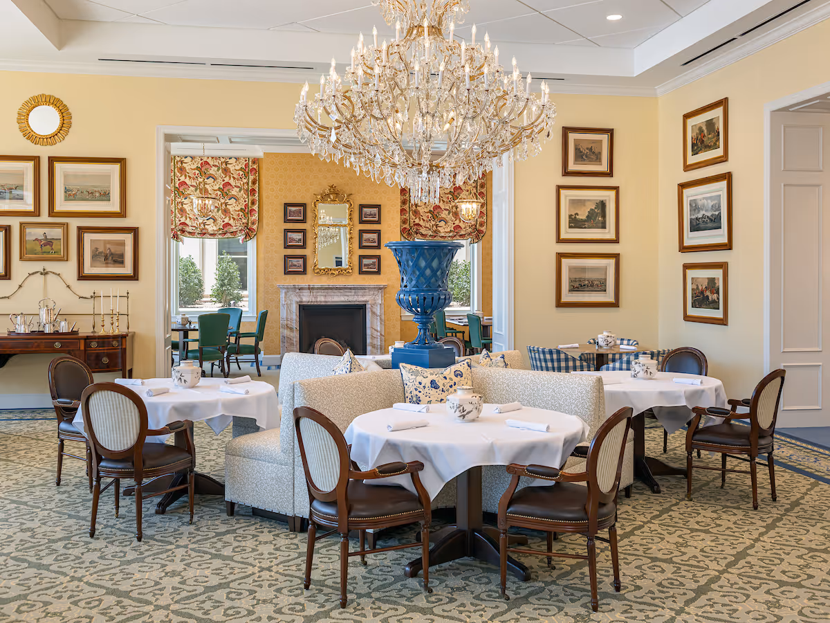 Elegant dining room with round tables set for service, upholstered seating, a large crystal chandelier, framed artwork, and a fireplace.
