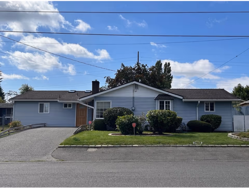 Single-story blue house with a front lawn, shrubs, and a driveway under a partly cloudy sky.