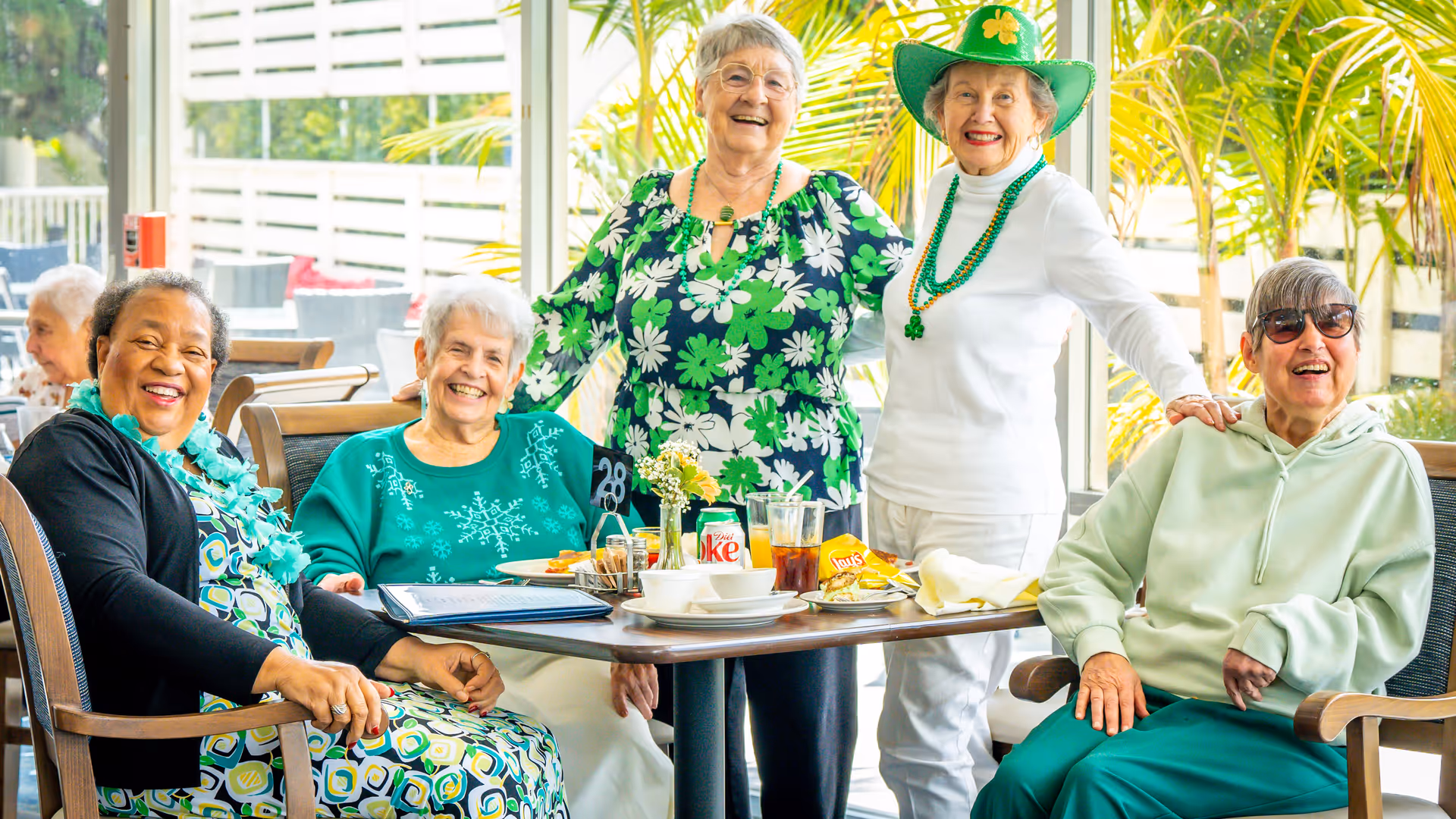 A group of five elderly women smiling and posing around a dining table in a bright room with large windows and green plants outside. The table has plates, drinks, and snacks, and some of the women are wearing green-themed clothing and accessories, suggesting a festive or themed gathering.