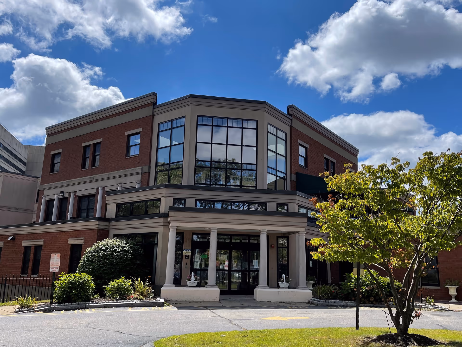 Front exterior of a three-story brick building with large glass windows, a columned entrance and a tree in the foreground under a partly cloudy sky.