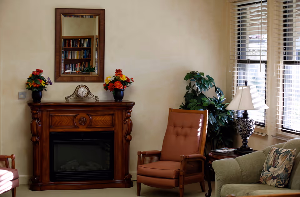 A cozy living room area with a wooden electric fireplace mantel decorated with a clock and two flower vases. Above the mantel is a mirror reflecting a bookshelf. To the right, there is a cushioned armchair, a side table with a decorative lamp, a leafy green plant, and a window with blinds letting in natural light. A green sofa with a floral pillow is partially visible.