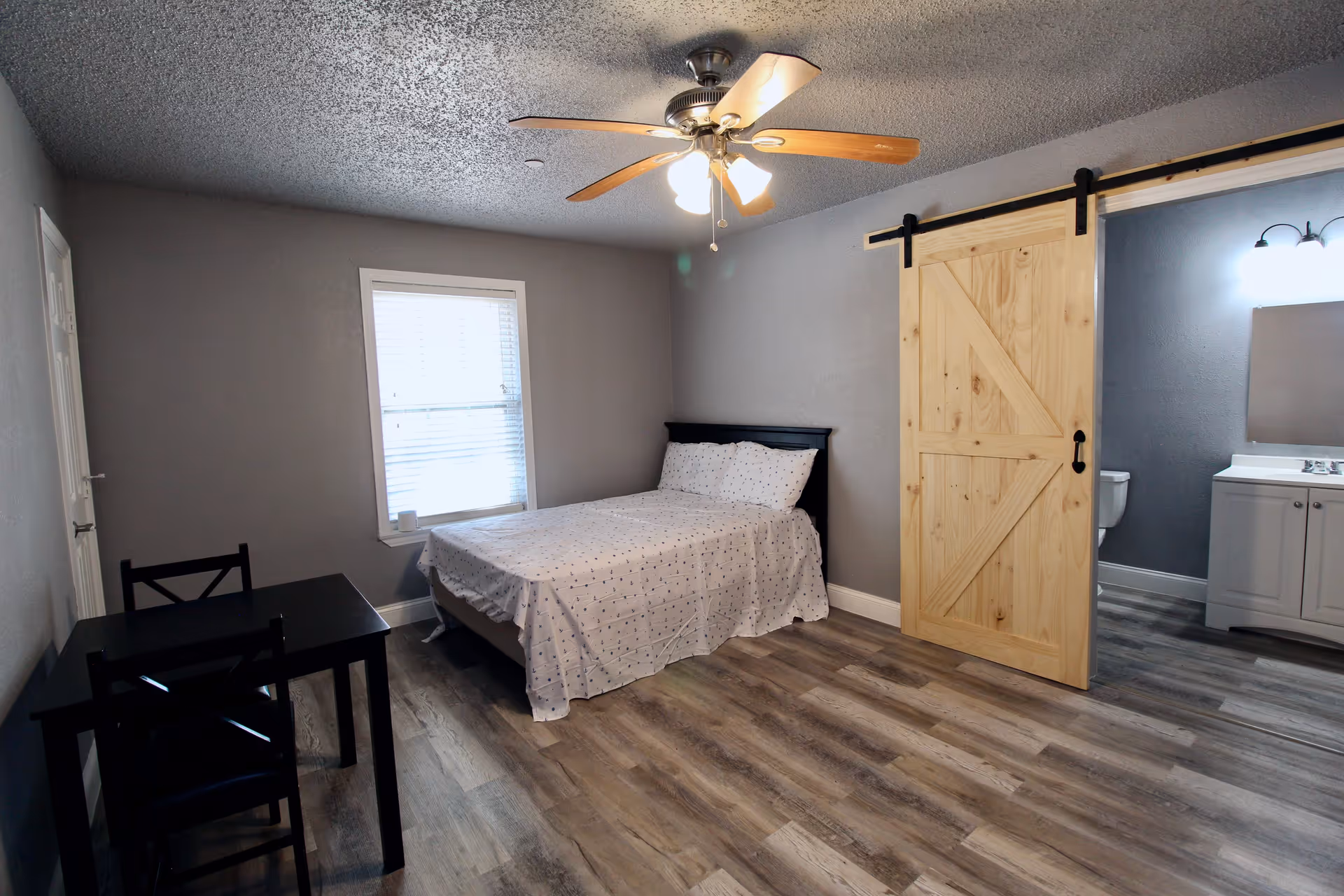 A furnished bedroom with a bed against gray walls, a ceiling fan, a small table and chairs, and a wooden sliding barn door opening to a bathroom.