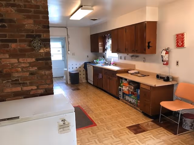 A kitchen area with wooden cabinets, a countertop, a dishwasher, and a sink. There is a brick wall on the left side, a white freezer in the foreground, and a door at the far end. A fire extinguisher and a fire safety sign are mounted on the wall to the right, next to an orange chair.