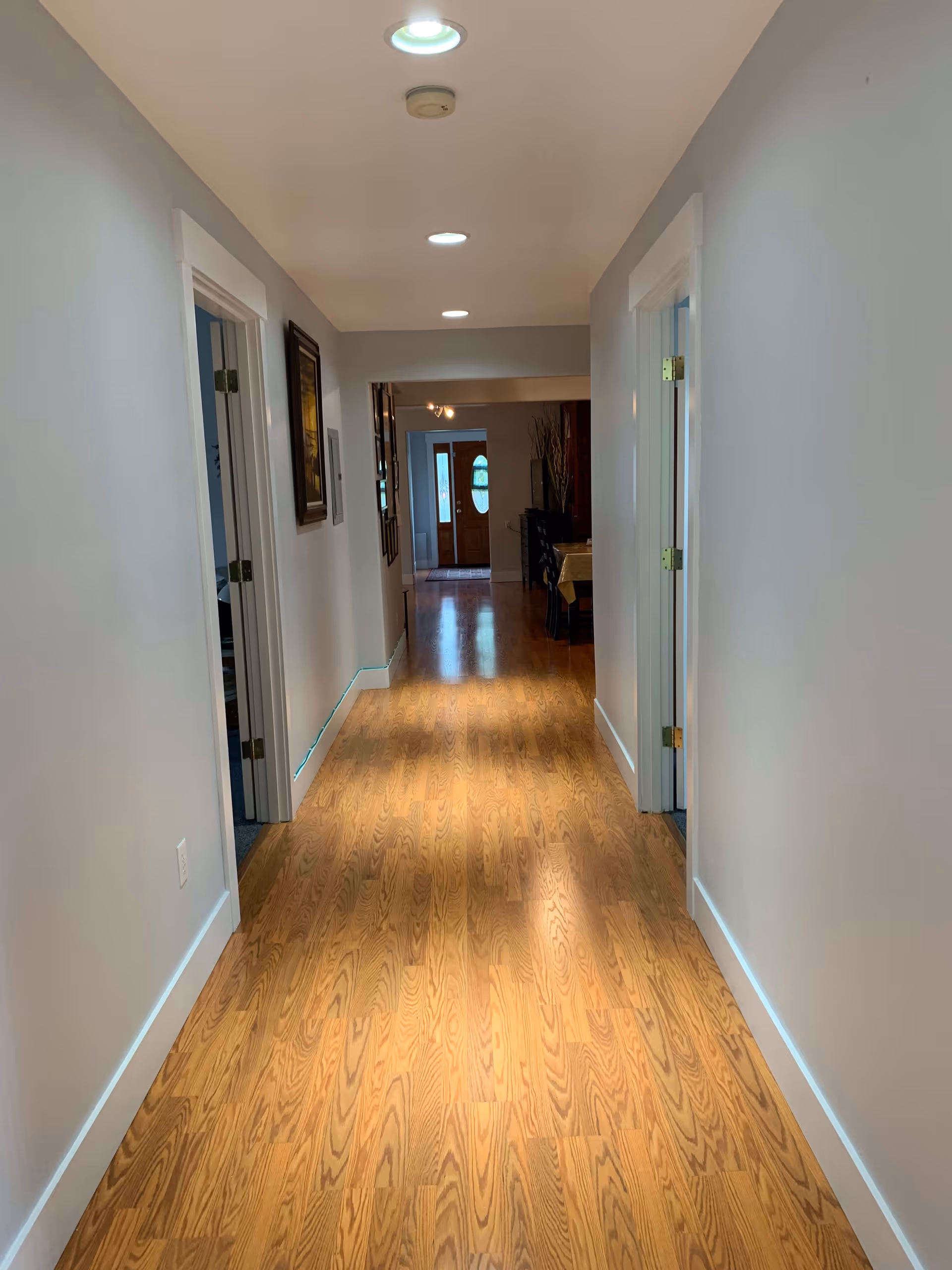 A long indoor hallway with wooden flooring and light gray walls. There are recessed ceiling lights and a smoke detector on the ceiling. Doors on both sides of the hallway lead to other rooms. At the end of the hallway, there is a dining area with a table and chairs, and a wooden front door with glass panels.