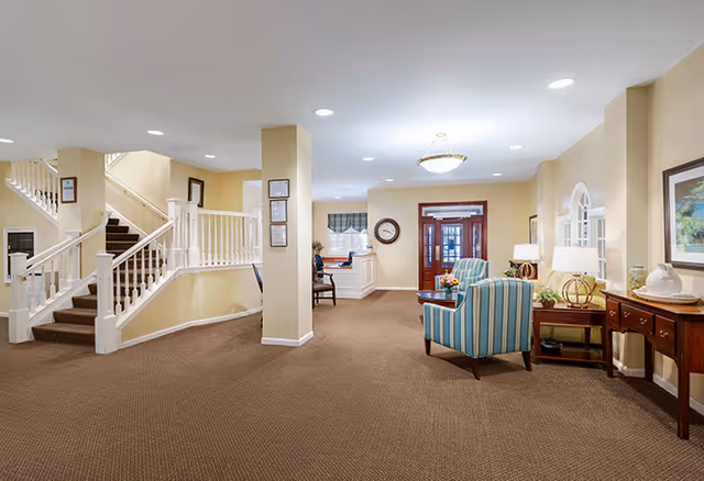 Spacious senior living facility lobby with seating area, striped armchairs, console tables, and a staircase leading to an upper level.