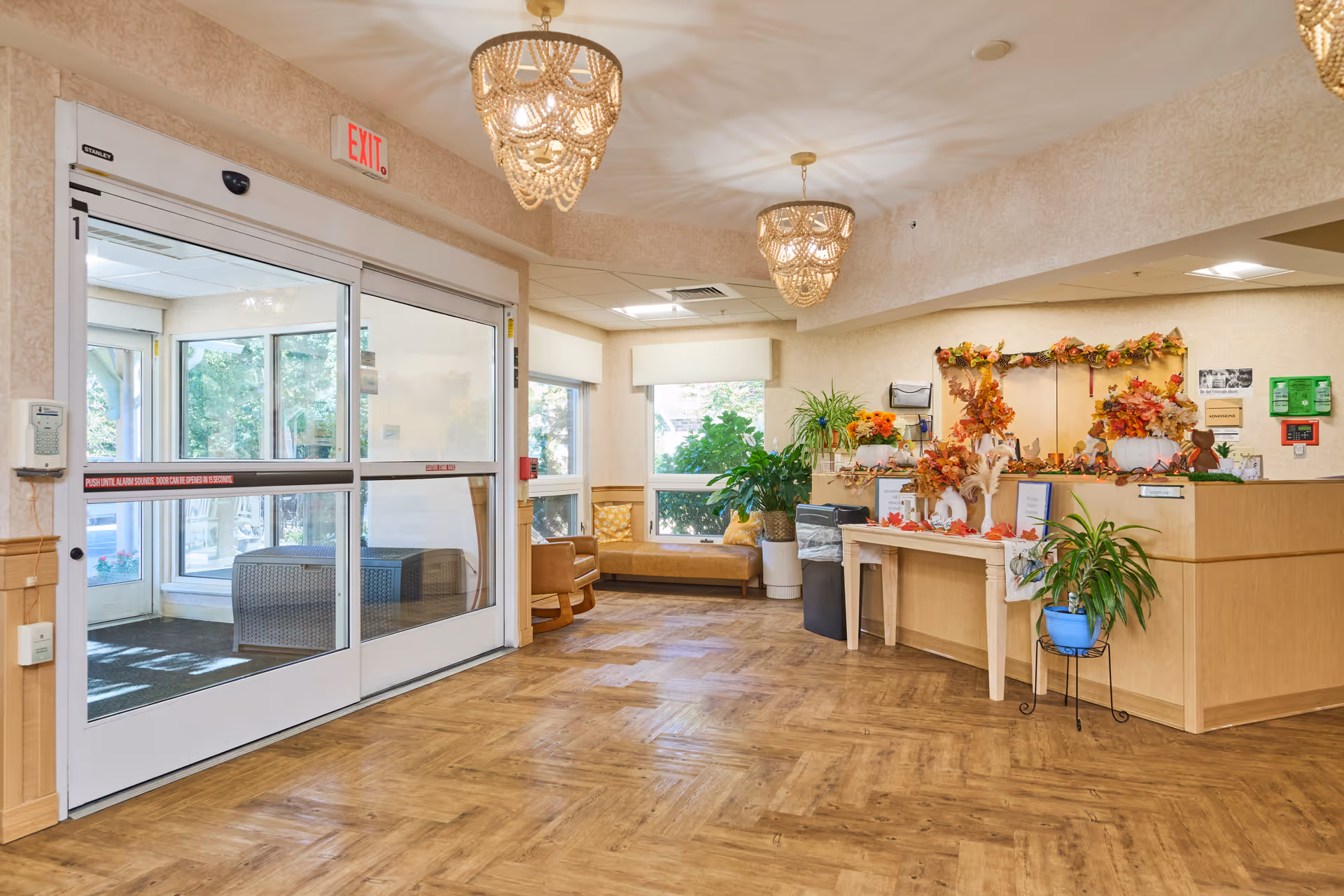 Interior view of a nursing facility entrance area with automatic sliding glass doors on the left, a reception desk decorated with autumn-themed decorations on the right, wooden flooring, two hanging chandeliers, and seating area with chairs and a bench near windows in the background.