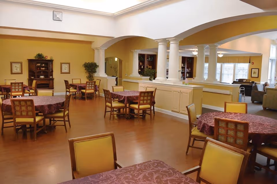 Interior view of a senior living facility dining room with several round tables covered with maroon tablecloths and surrounded by wooden chairs with yellow cushions. The room has yellow walls, wooden flooring, white columns, and an open archway leading to a lounge area with sofas and large windows.