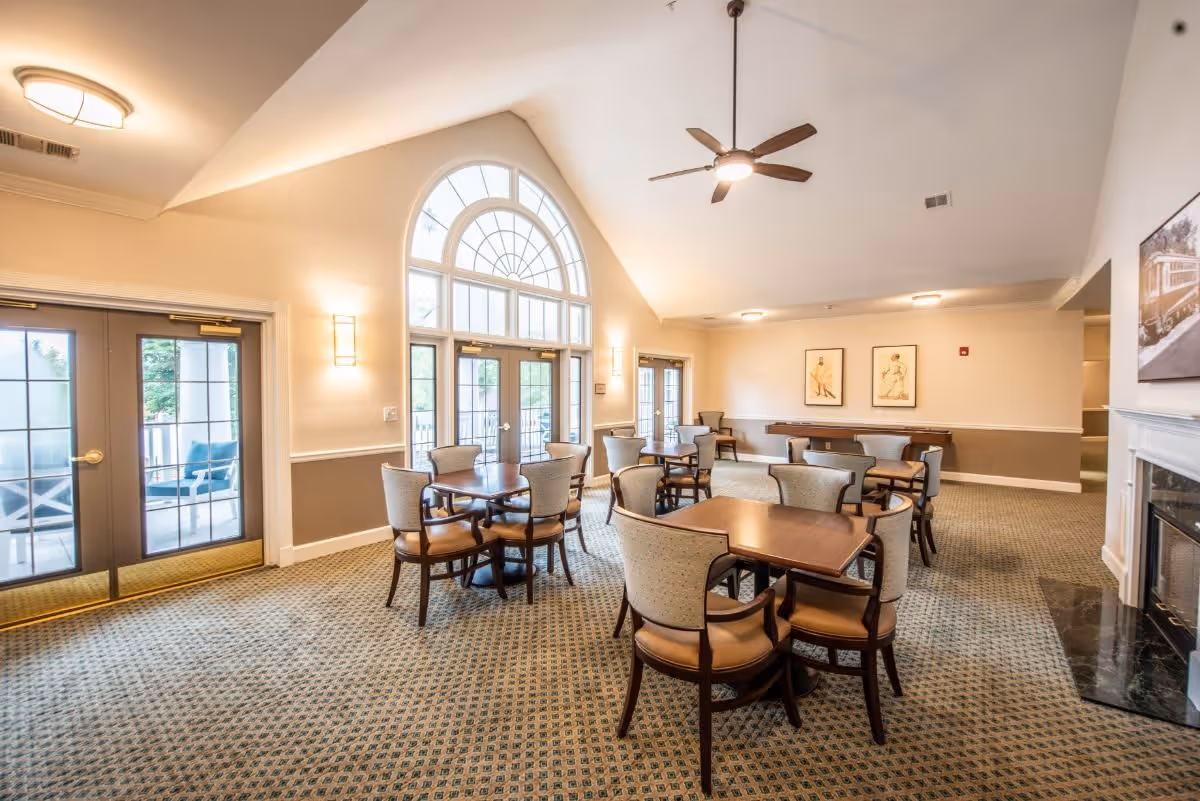 A spacious, well-lit common area with multiple tables and chairs arranged for seating. The room features large arched windows and glass doors allowing natural light to enter. The walls are painted in neutral tones with framed artwork, and there is a ceiling fan and a fireplace on one side. The carpet has a patterned design, and the overall atmosphere is inviting and comfortable.