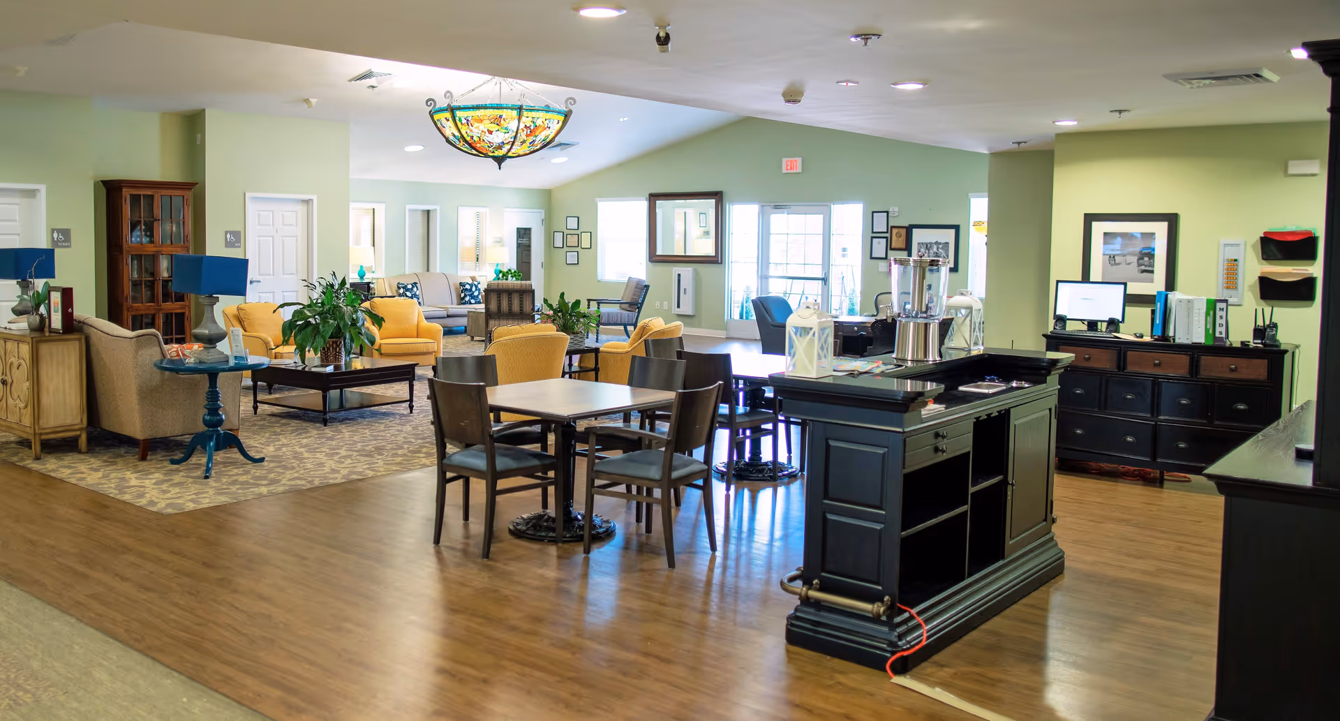 Spacious senior living common area with seating clusters, dining tables, and a central service island under a decorative ceiling light.