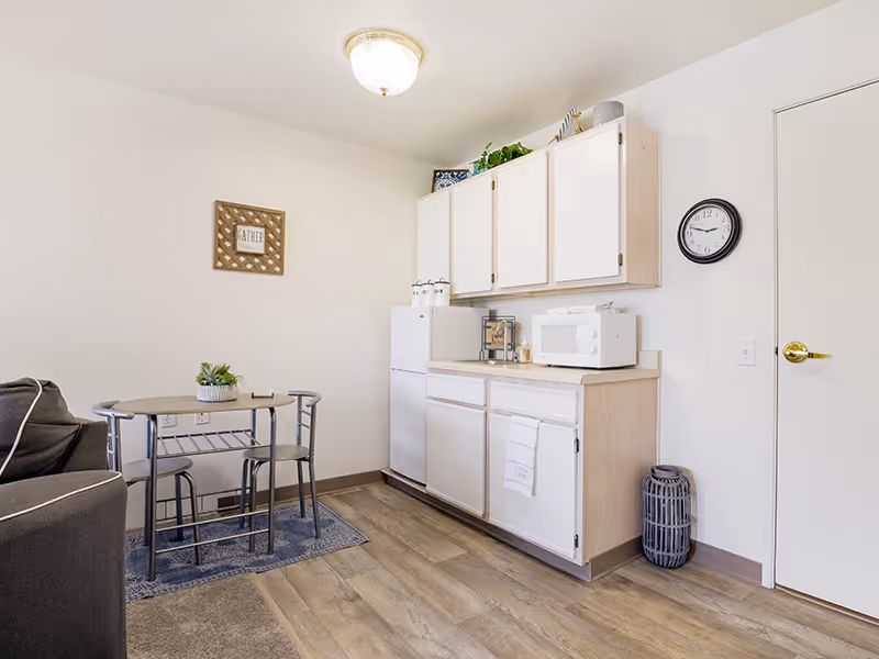 Small studio kitchenette with white cabinets, a refrigerator and microwave, a small dining table and a couch on wood-look flooring.