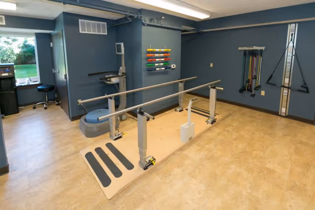Rehabilitation room with parallel walking bars, exercise equipment, resistance bands, and a small seated area by a window.