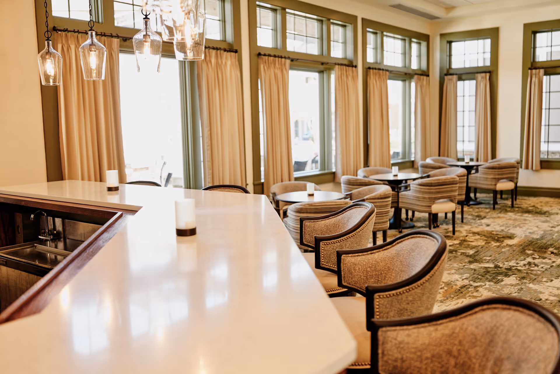 Bright dining and lounge area with a long marble counter, upholstered chairs, small round tables, and large windows with curtains.