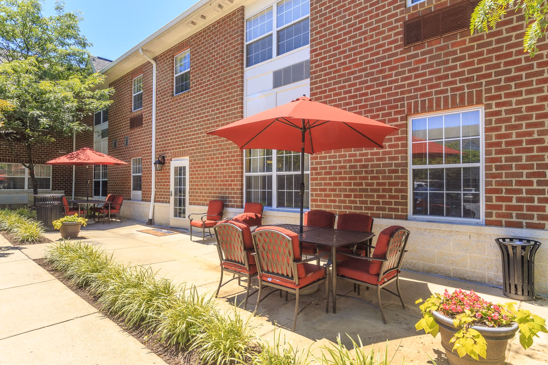 Outdoor patio area at Arbor Crest of Silver Spring with red cushioned chairs around tables shaded by red umbrellas, adjacent to a brick building with windows and a door. There are plants and greenery along the sidewalk.