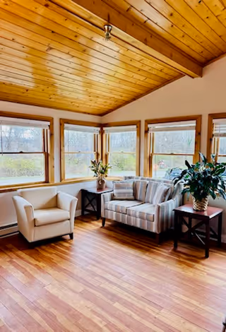 Sunlit sitting area with a wood-paneled vaulted ceiling, hardwood floor, a striped loveseat, an armchair, side tables and large multi-pane windows.