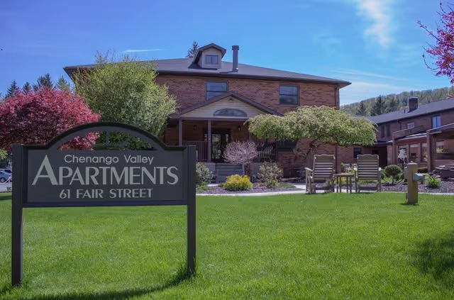 Exterior view of Chenango Valley Apartments building with a green lawn and outdoor seating area. A sign in the foreground reads 'Chenango Valley Apartments 61 Fair Street'. Trees and shrubs surround the building under a clear blue sky.