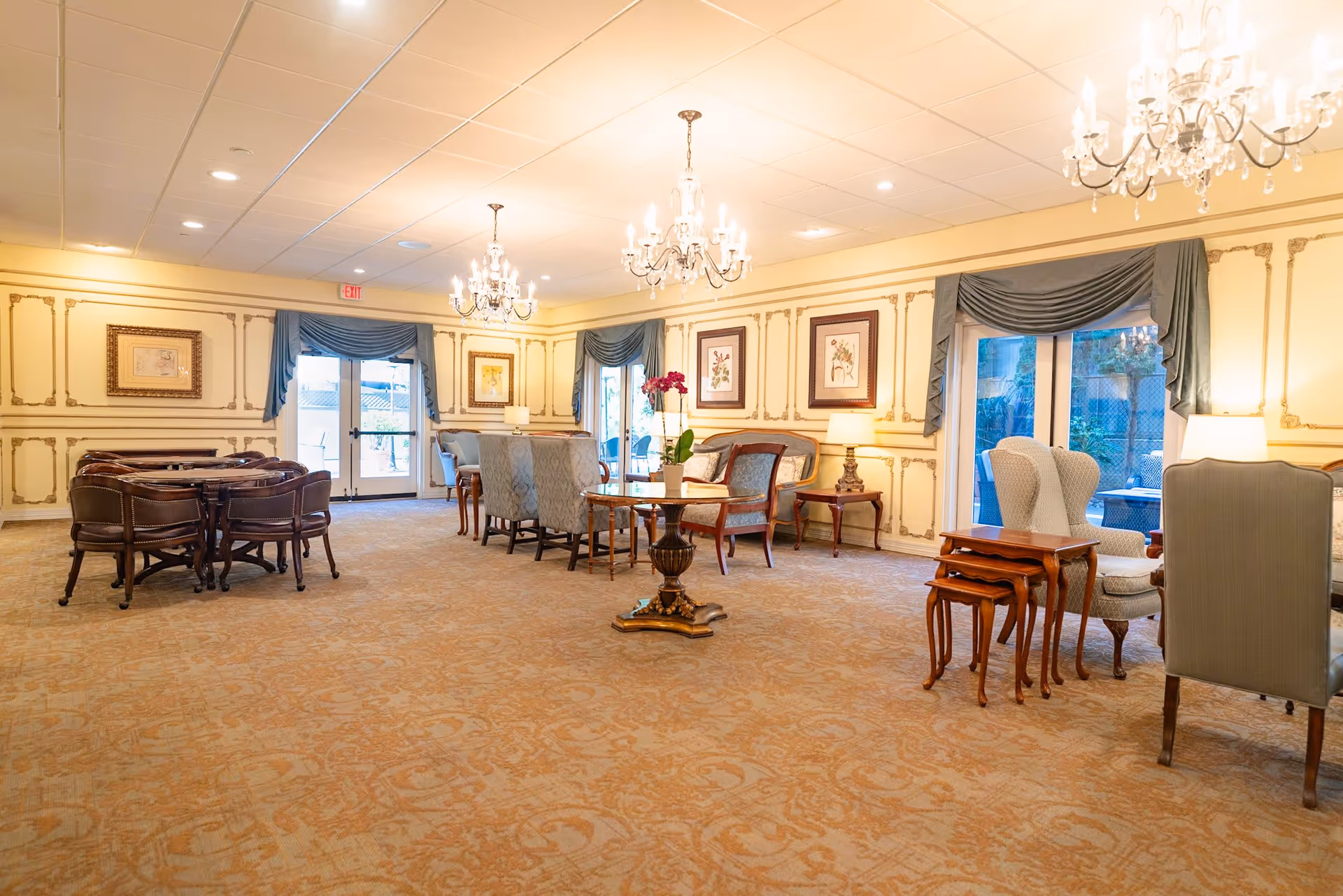A spacious, elegantly decorated common area with multiple seating arrangements including upholstered chairs and wooden tables. The room features ornate wall paneling, framed artwork, large windows with blue drapes, and three crystal chandeliers hanging from the ceiling. The carpet has a subtle patterned design, and there are table lamps providing additional lighting.
