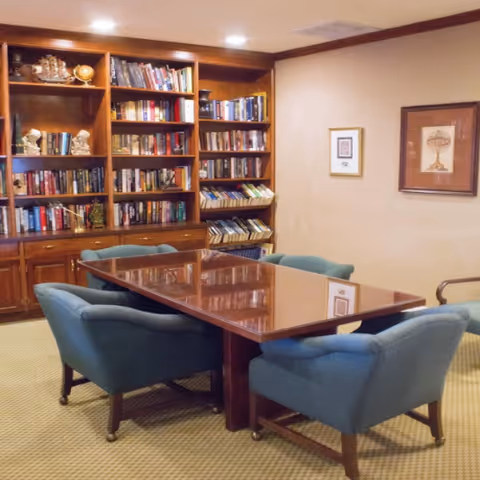 A cozy room with a wooden table surrounded by six blue upholstered chairs on casters. Behind the table is a large wooden bookshelf filled with books and decorative items. The walls are beige with two framed pictures hanging on the right side.