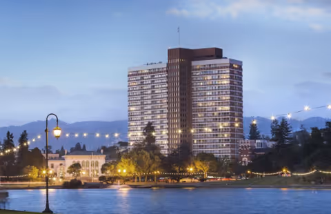 A tall multi-story residential building named St. Paul's Towers illuminated at dusk, with string lights and street lamps along the waterfront in the foreground and hills in the background.