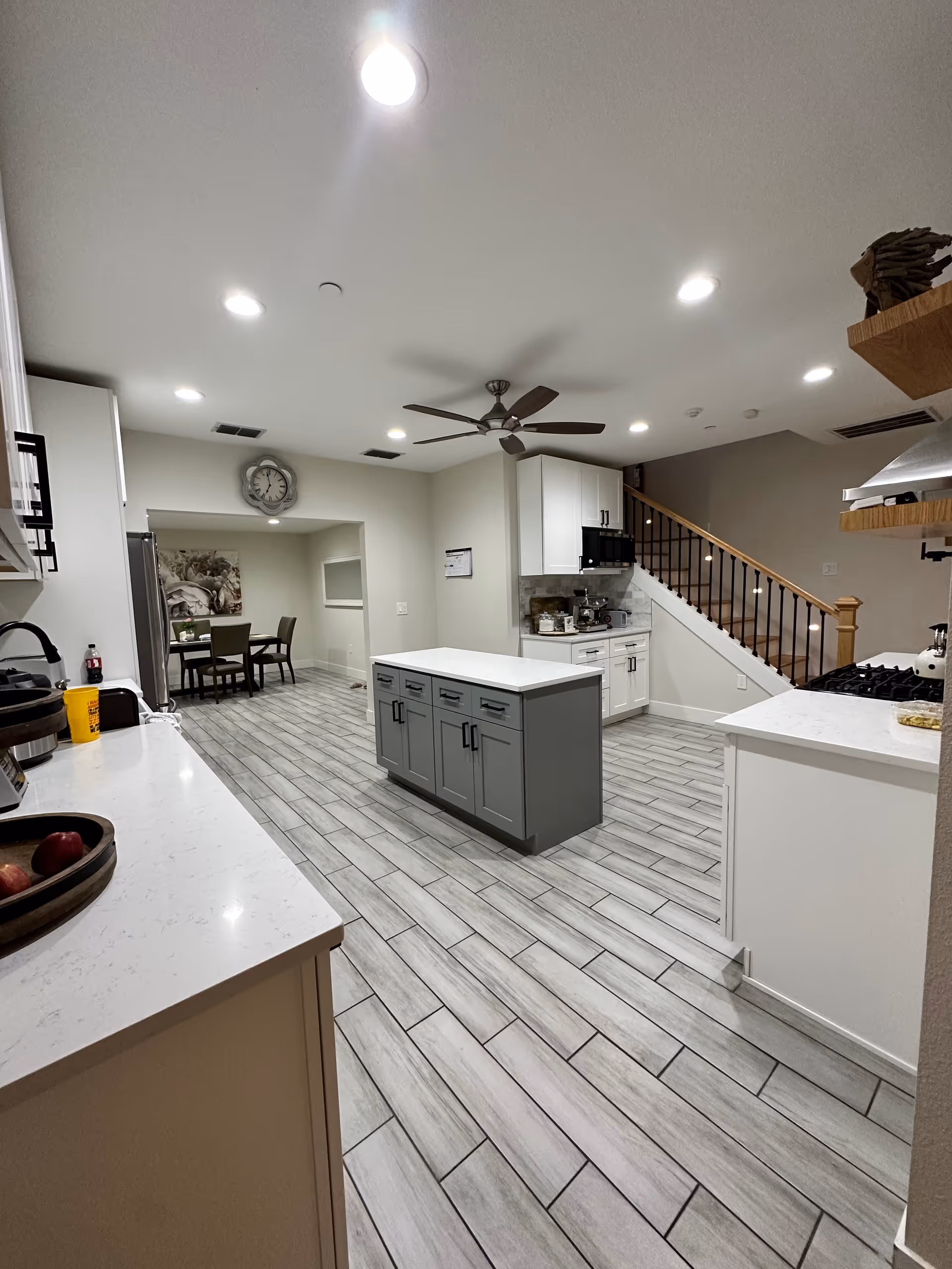 A modern kitchen with light gray tiled flooring, white cabinets, and a central island with gray cabinetry. The kitchen features a ceiling fan, recessed lighting, a stove with a range hood, and a dining area with a table and chairs visible through an open doorway. There is a staircase with wooden handrails and black balusters on the right side.
