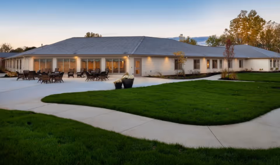 Exterior view of a single-story building with a gray roof and beige siding, surrounded by a well-maintained lawn and concrete walkways. Several outdoor tables and chairs are arranged on a patio area near large windows. Trees with autumn foliage are visible in the background under a clear sky at dusk.