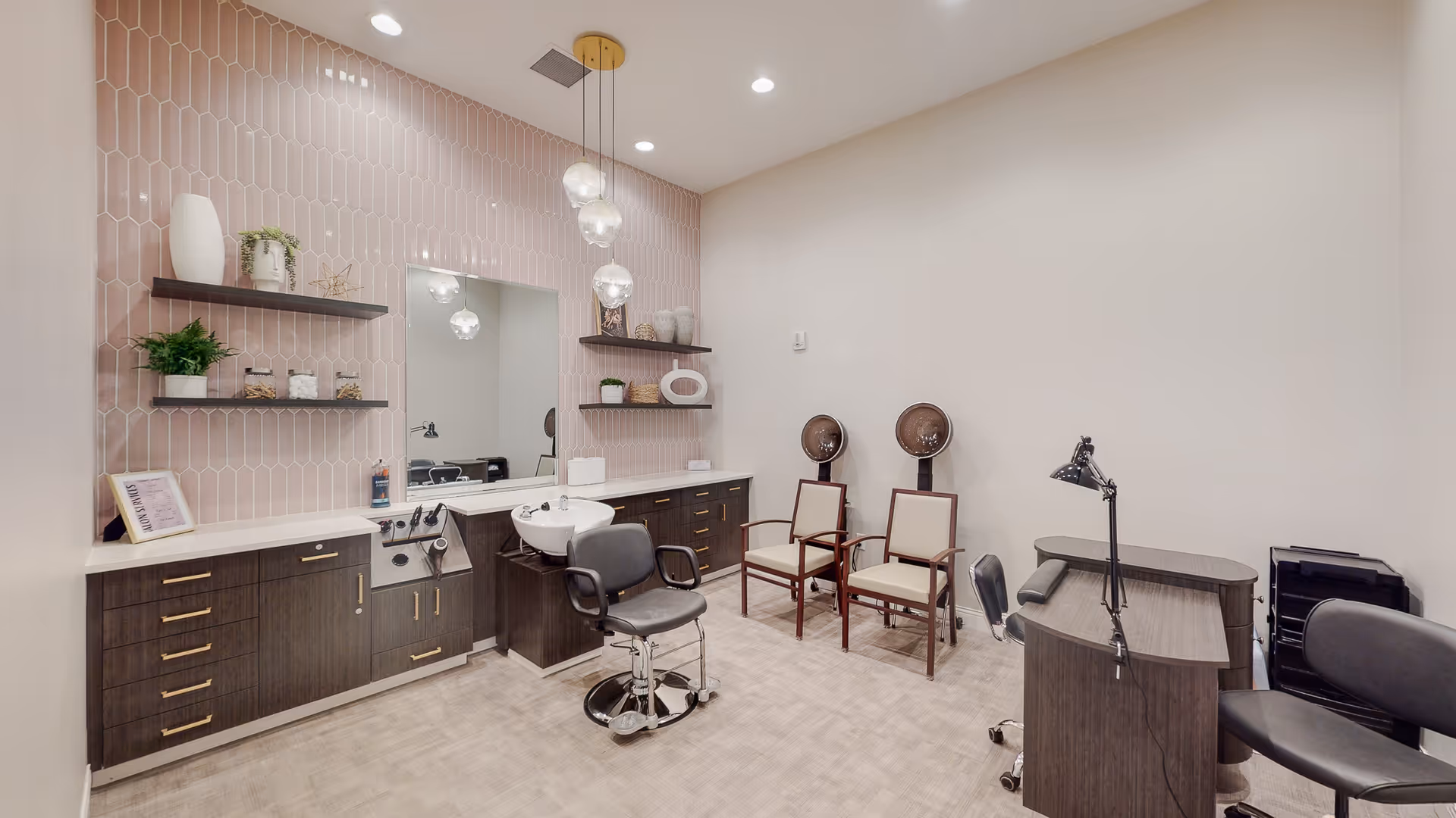 Interior of a salon room with a styling chair in front of a sink, two hair drying chairs with hooded dryers, a manicure station with a lamp, and cabinets with shelves decorated with plants and ornaments against a pink tiled wall.
