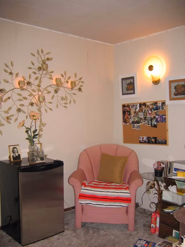 A cozy corner of a room featuring a pink upholstered armchair with a striped blanket and a brown pillow. Next to the chair is a small stainless steel mini fridge with framed photos and a vase with flowers on top. The wall behind has a decorative metal wall art shaped like a tree with flowers, a bulletin board filled with photos, and two framed pictures. A round glass side table holds various items including books and tissues. The room has beige walls and carpeted floor.